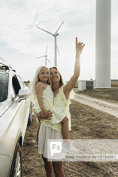 Smiling woman gesturing and standing with daughter by car at wind park