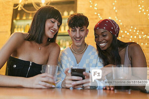 Happy young man sharing smart phone sitting amidst friends in restaurant