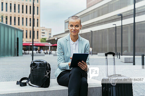 Smiling businesswoman with tablet PC and luggage sitting on bench at footpath