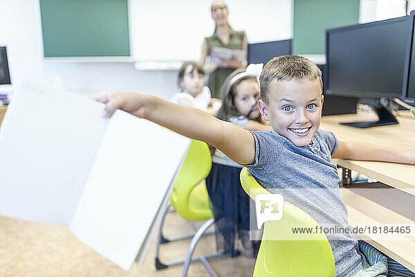 Excited boy showing notebook sitting on chair in classroom