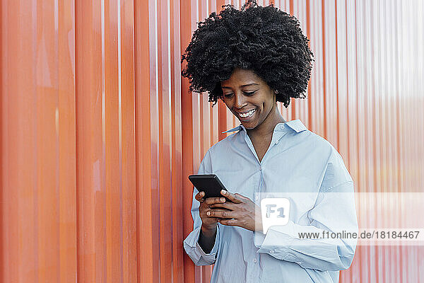Smiling young woman using smart phone standing by orange wall