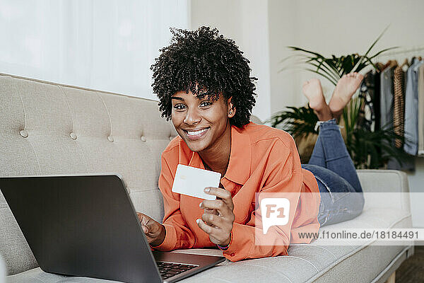 Happy freelancer lying on sofa with laptop and credit card at home office
