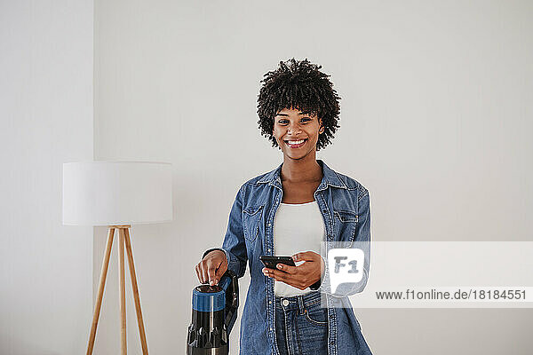 Happy woman standing with vacuum cleaner and smart phone in front of wall at home