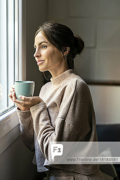 Thoughtful woman standing with cup of tea near window at home