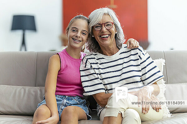 Happy girl sitting with arm around by grandmother on sofa at home
