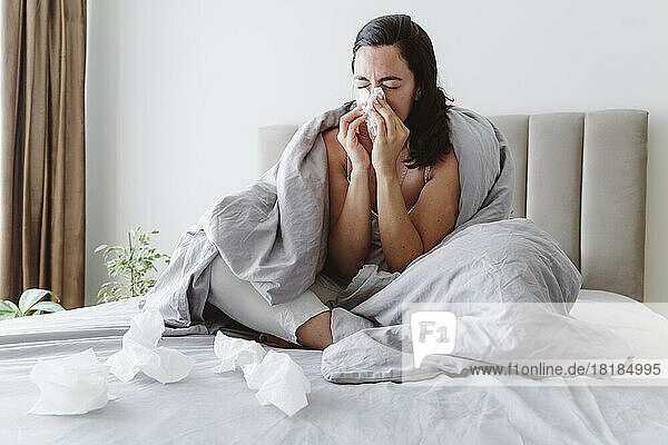 Ill woman with eyes closed blowing nose in tissue on bed at home