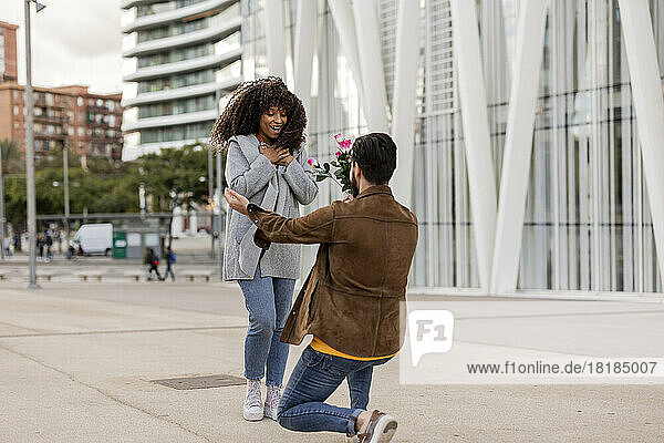 Junger Mann macht Frau mit Blumenstrauß am Fußweg einen Heiratsantrag