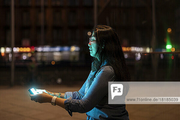 Mature woman wearing smart glasses using smart phone on footpath at night