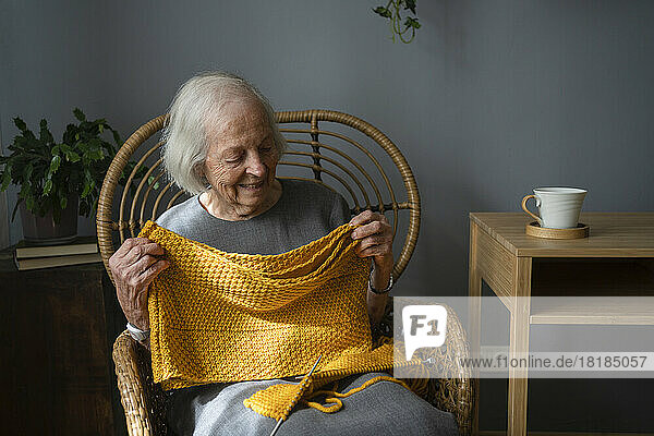 Happy senior woman holding knitted scarf on chair at home
