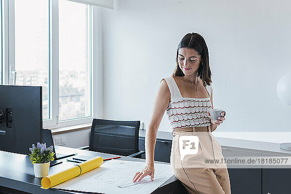 Young architect looking at blueprint holding coffee cup in office