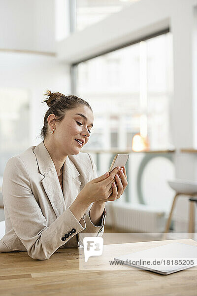 Businesswoman looking at mobile phone sitting with laptop at desk