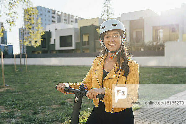 Smiling young woman wearing helmet standing with push scooter in park on sunny day