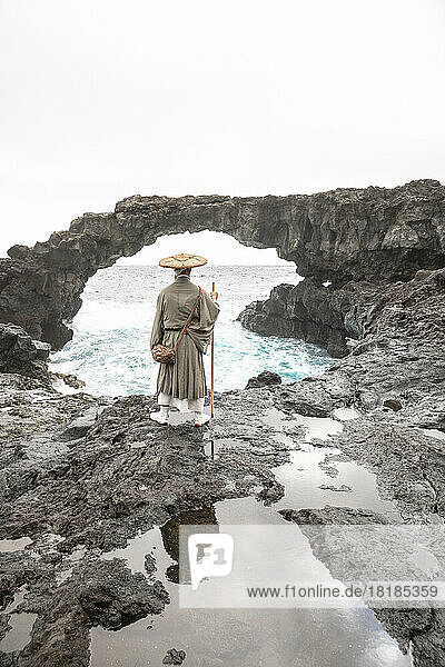 Mature monk with stick standing in front of rock arch