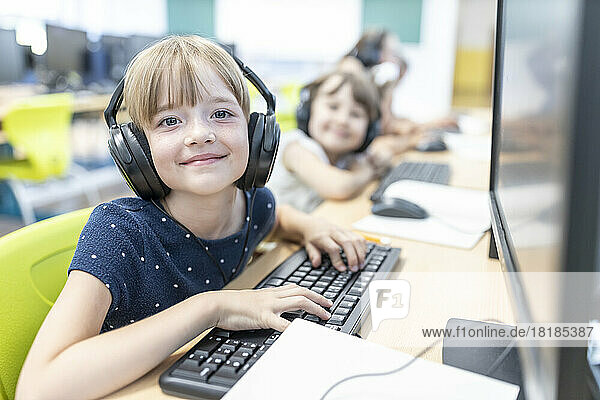 Smiling girl with bangs wearing headphones sitting at desk by computer in class