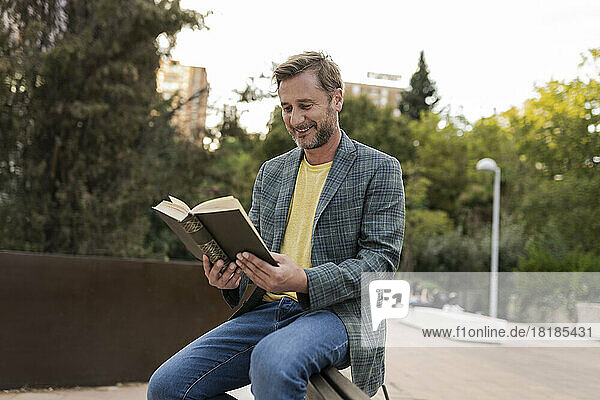 Happy man reading book sitting on bench at park