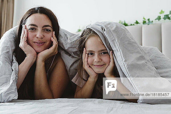 Smiling woman and daughter with head in hands under blanket lying on bed
