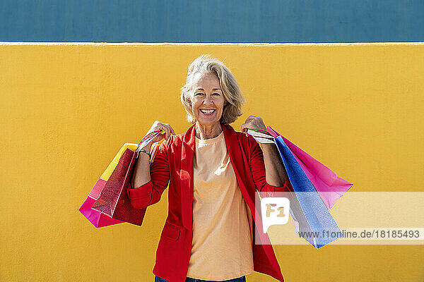 Happy mature woman with multi colored shopping bags in front of yellow wall
