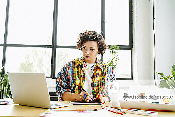 Young graphic designer using laptop sitting in front of window at desk