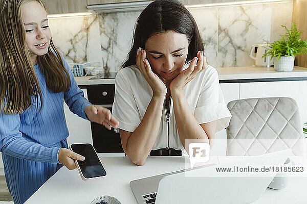 Girl holding mobile phone looking at stressed mother sitting with laptop on table