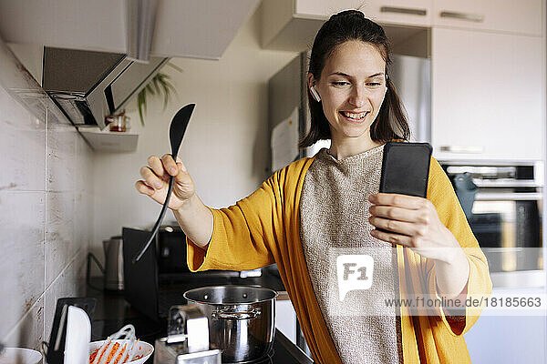 Smiling young woman holding wooden spoon having video call through mobile phone in kitchen
