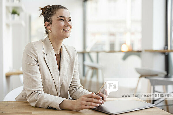 Smiling businesswoman holding mobile phone sitting with laptop at desk