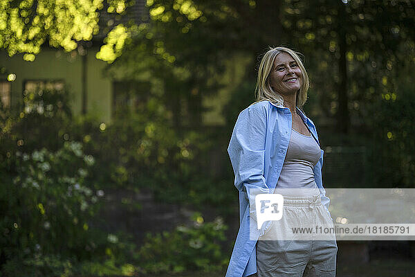Happy mature woman standing with hands in pockets at garden