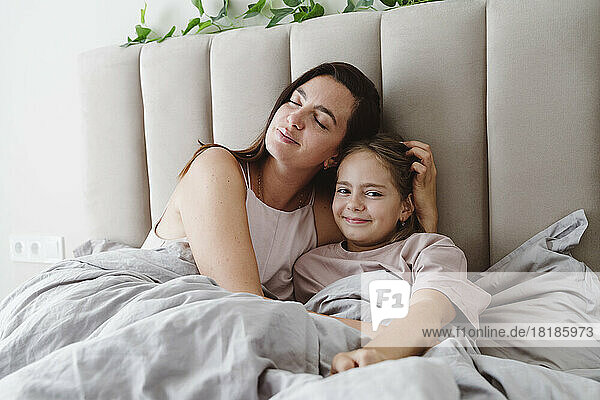 Woman with eyes closed embracing daughter lying on bed at home