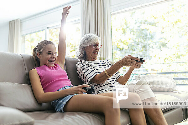 Granddaughter with hand raised playing video game with grandmother sitting on sofa at home