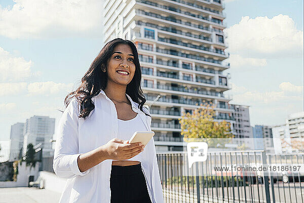 Smiling young woman with smart phone standing in front of building