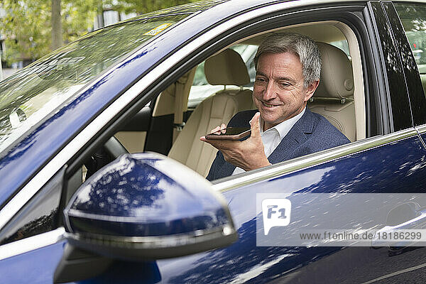 Smiling mature businessman sitting in car talking on speaker phone