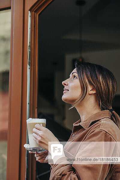 Thoughtful young woman with glass of cappuccino standing in doorway at cafe