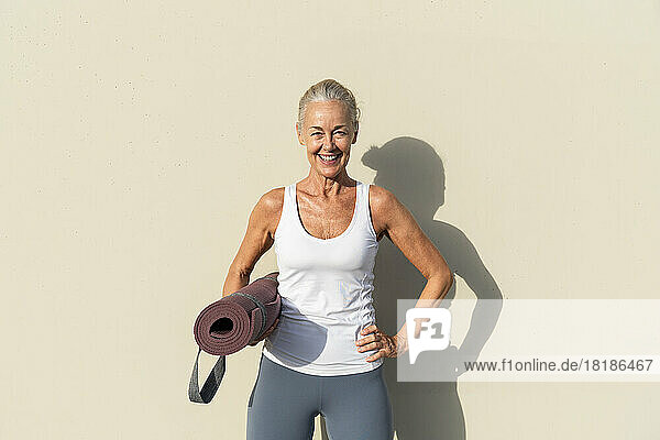 Smiling woman with yoga mat in front of cream colored wall