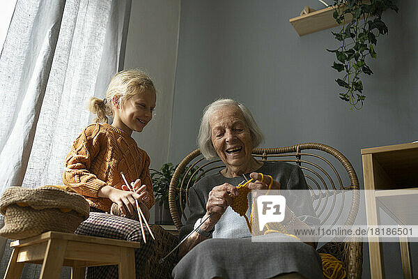 Happy girl holding ball of wool and needle looking at grandmother knitting on chair