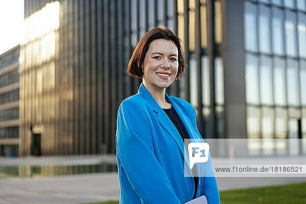 Lächelnde junge Geschäftsfrau im blauen Blazer steht vor dem Gebäude