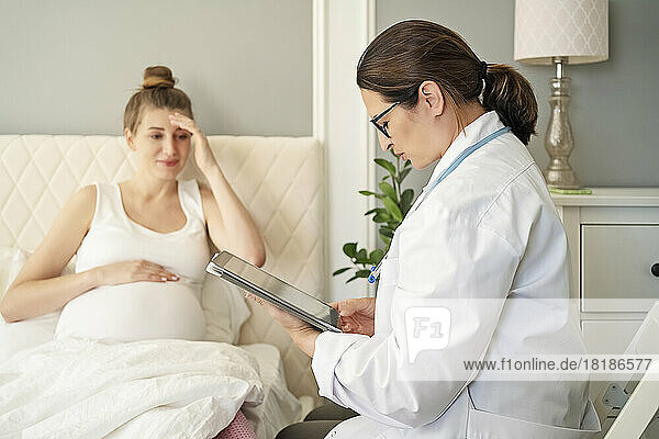 Doctor with tablet PC consulting pregnant woman on bed at home