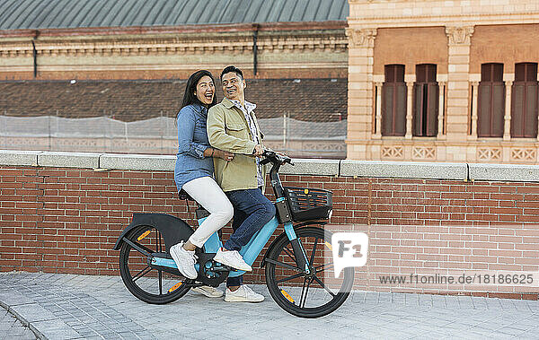 Happy mature couple sitting on electric bicycle by brick wall