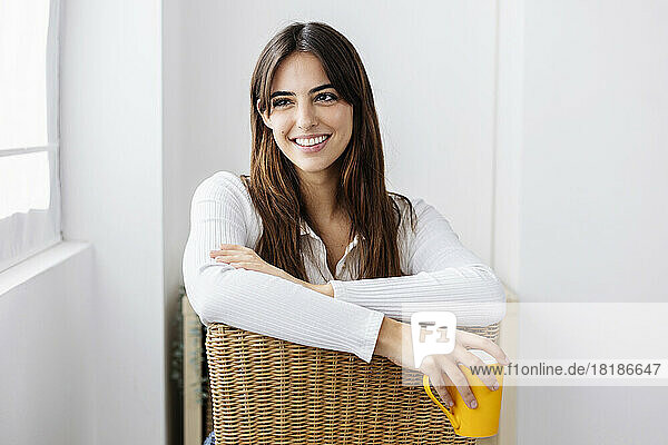 Lächelnde Frau mit Kaffeetasse sitzt zu Hause auf einem Stuhl