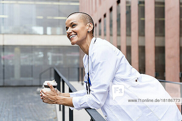 Lächelnder Arzt mit Einweg-Kaffeetasse lehnt am Geländer