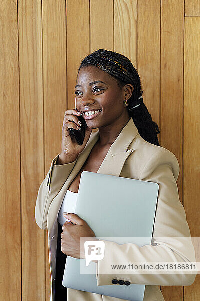 Happy young businesswoman with laptop talking on smart phone in front of wooden wall