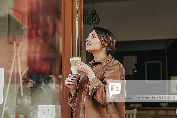 Nachdenkliche Frau mit einem Glas Cappuccino steht in der Tür eines Cafés