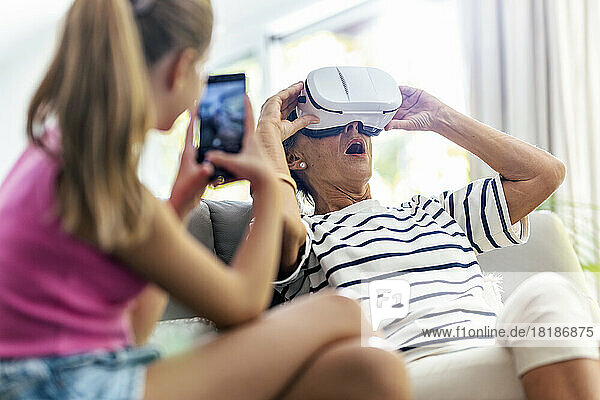 Girl photographing grandmother wearing virtual reality headset at home