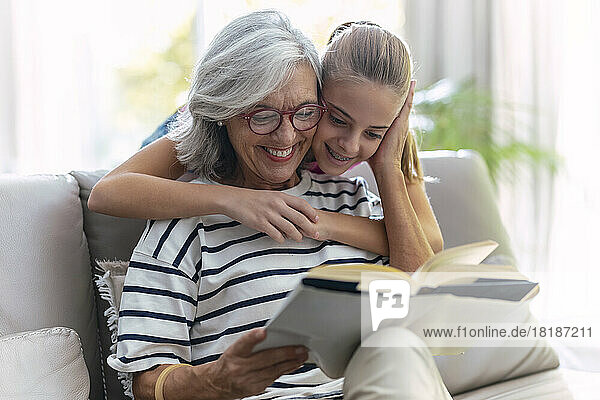 Lächelndes Mädchen umarmt Großmutter und liest zu Hause auf dem Sofa ein Buch