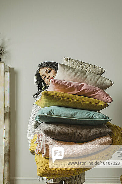 Smiling woman holding pillows and blankets in front of wall at home