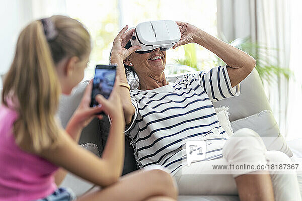 Granddaughter photographing senior woman enjoying virtual reality headset at home