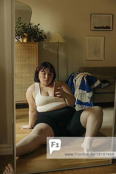 Volle Länge der jungen üppigen Frau  die selfie durch smart phone beim Sitzen gegen Spiegel zu Hause