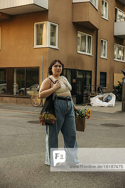 Portrait of confident young woman with shopping bag standing against building