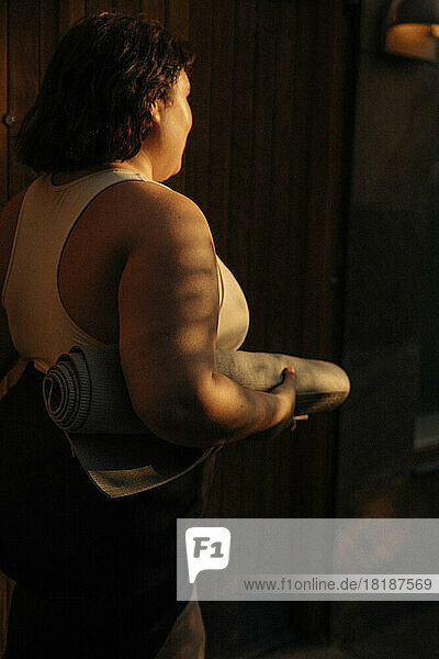Seitenansicht einer jungen Frau mit Trainingsmatte  die bei Sonnenuntergang im Hinterhof spazieren geht
