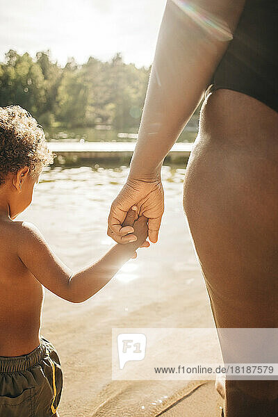 Frau hält die Hand ihres Sohnes  während sie an einem sonnigen Tag in der Nähe eines Sees steht