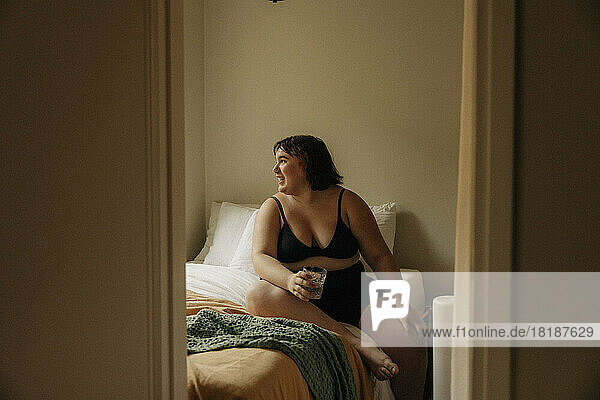 Volle Länge der glücklichen jungen Frau in Dessous sitzt mit Trinkglas im Schlafzimmer