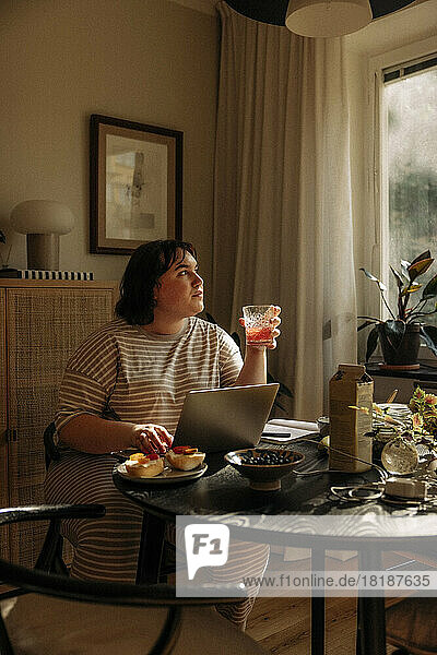 Junge Frau hält ein Trinkglas in der Hand und sitzt mit einem Laptop am Tisch